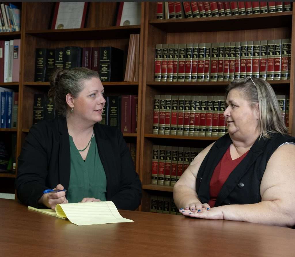 Kristen Howe and Cheri Mitchell from the Georgia Advocacy Office are sitting at a wooden table in a room filled with bookshelves. Kristen appears to be speaking to Cheri on the right, who is listening attentively. Both are dressed in professional attire.